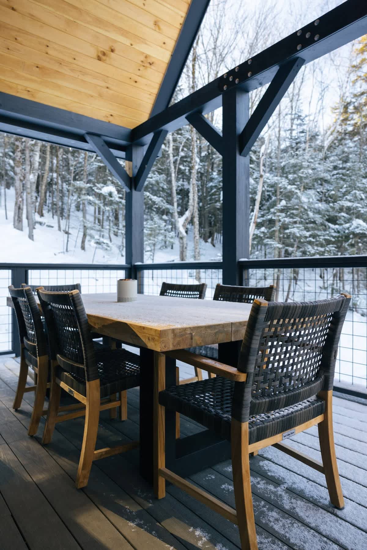 A covered outdoor deck features a rustic wooden dining table surrounded by black woven chairs, set against a backdrop of a snow-covered forest. The deck is supported by dark beams and has black railings.