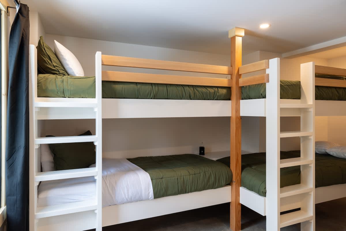 A bright bedroom features two sets of modern white and light wood bunk beds, each with olive green bedding and white pillows. A dark curtain is visible on the left side of the room.