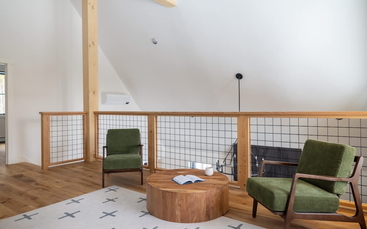 Loft lounge area with two green armchairs and a round wooden coffee table on a patterned rug. A modern railing with wire mesh overlooks the space below, and a wooden beam crosses the vaulted ceiling.