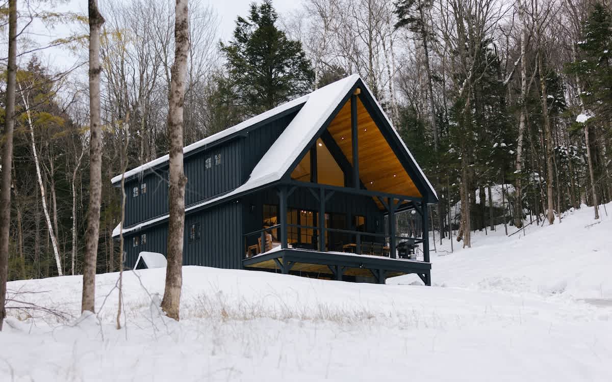 Modern A-frame cabin with dark siding and a snow-covered roof, nestled in a snowy, wooded landscape. A spacious deck with outdoor seating is visible, offering views of the surrounding forest.