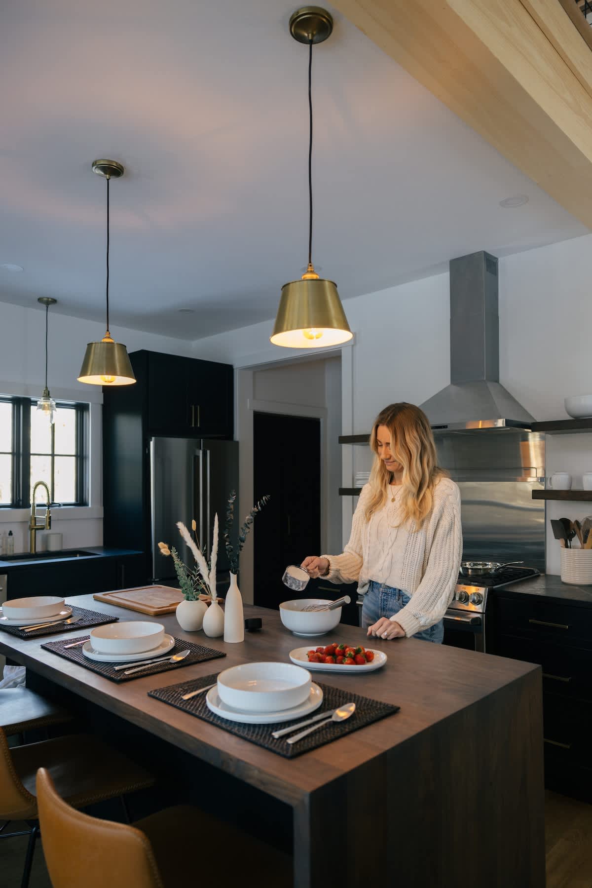 A woman adds ingredients to a bowl on a dark wood kitchen island, set with white dinnerware and black placemats. The kitchen features modern black cabinets, brass pendant lights, and stainless steel appliances.