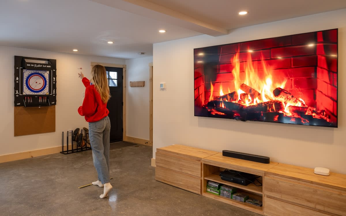 A woman plays darts in a spacious game room with a large wall-mounted TV displaying a digital fireplace. The room has a concrete floor, recessed lighting, and a dartboard setup.