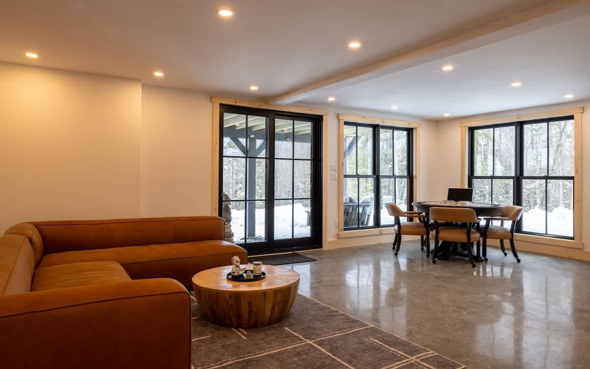A spacious living area features polished concrete floors, an L-shaped orange sectional sofa, and a round wooden coffee table on a patterned rug. Large black-framed windows and a glass door provide views of a snowy outdoor landscape.