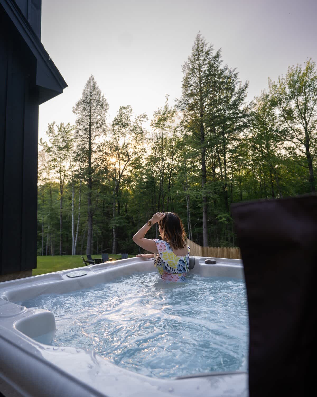 Outdoor hot tub surrounded by lush green trees under a soft evening sky, with a person relaxing in the bubbling water, enjoying the serene natural setting.