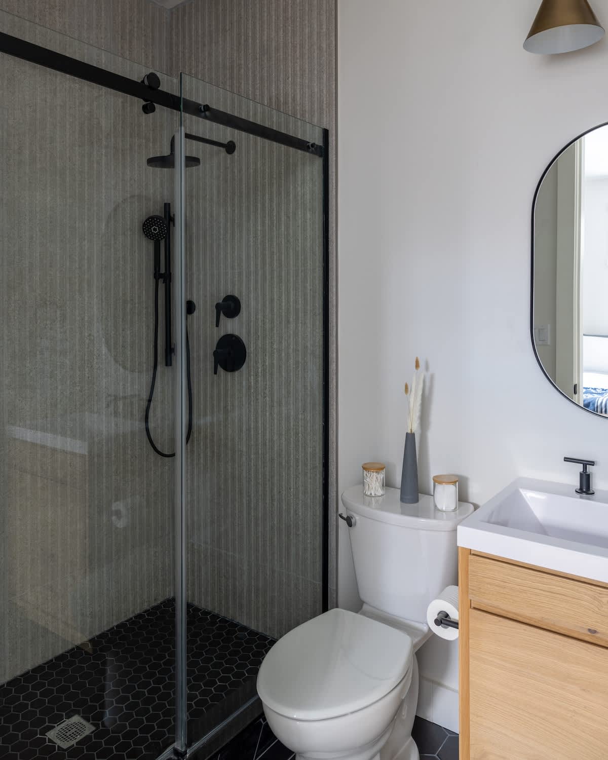 Modern bathroom featuring a walk-in shower with black fixtures and a sliding glass door, a white toilet, and a light wood vanity with a white sink and oval mirror. The shower has textured gray tile walls and a black hexagonal tile floor.
