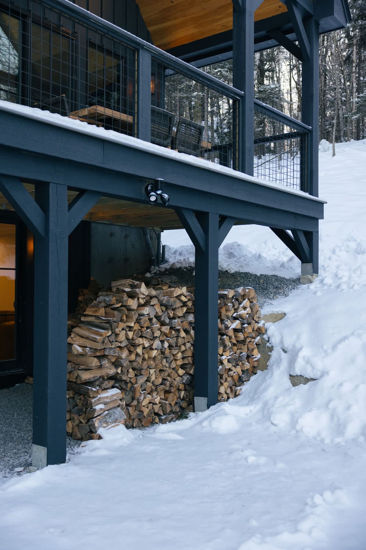 Exterior view of a dark-stained wooden deck supported by sturdy posts, with a large stack of firewood neatly stored underneath. The ground is covered in snow, indicating a winter setting.
