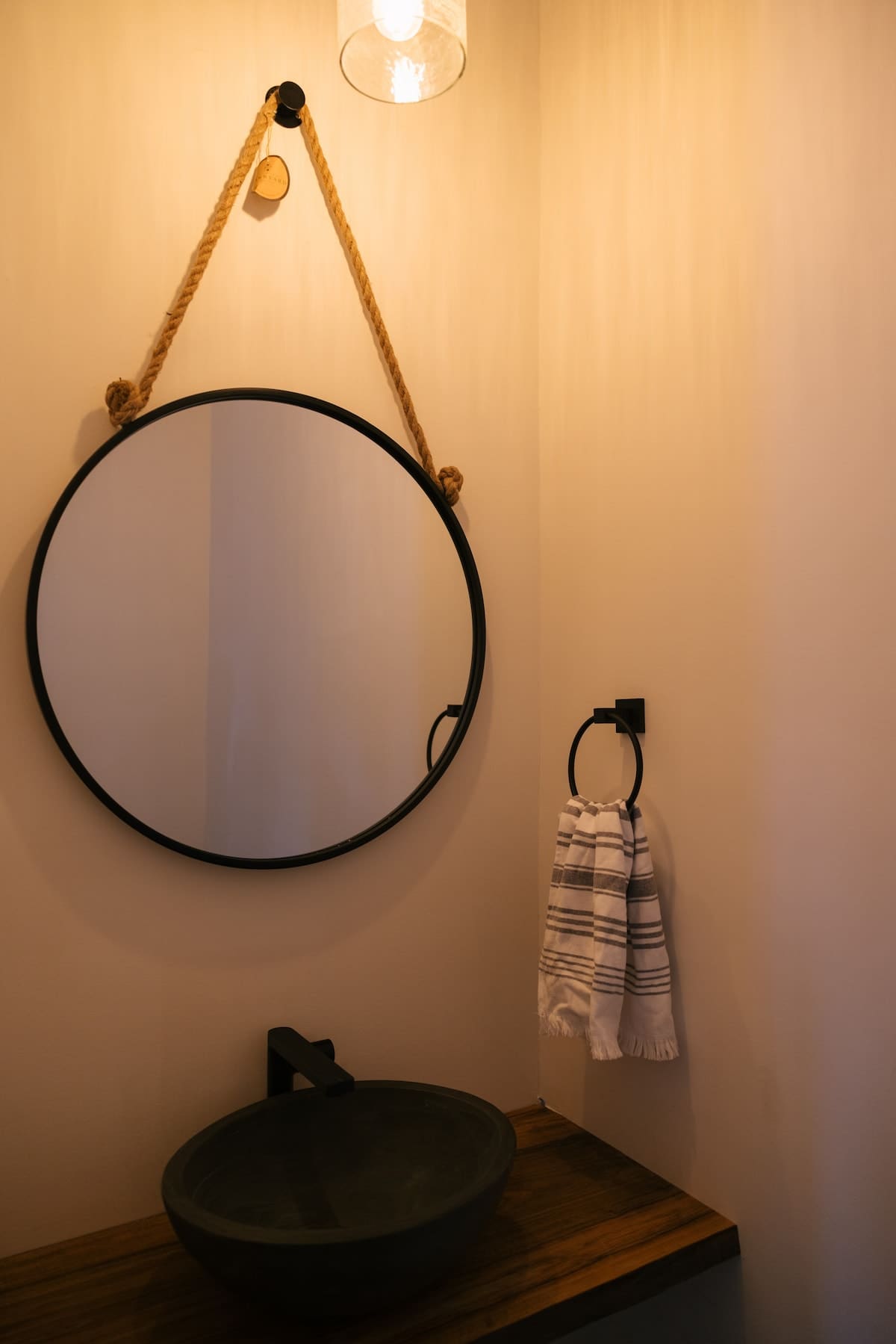 Cozy powder room featuring a round black-framed mirror suspended by rope, a dark vessel sink on a wooden countertop, and a black towel ring with a striped hand towel. Warm lighting illuminates the light-colored walls.