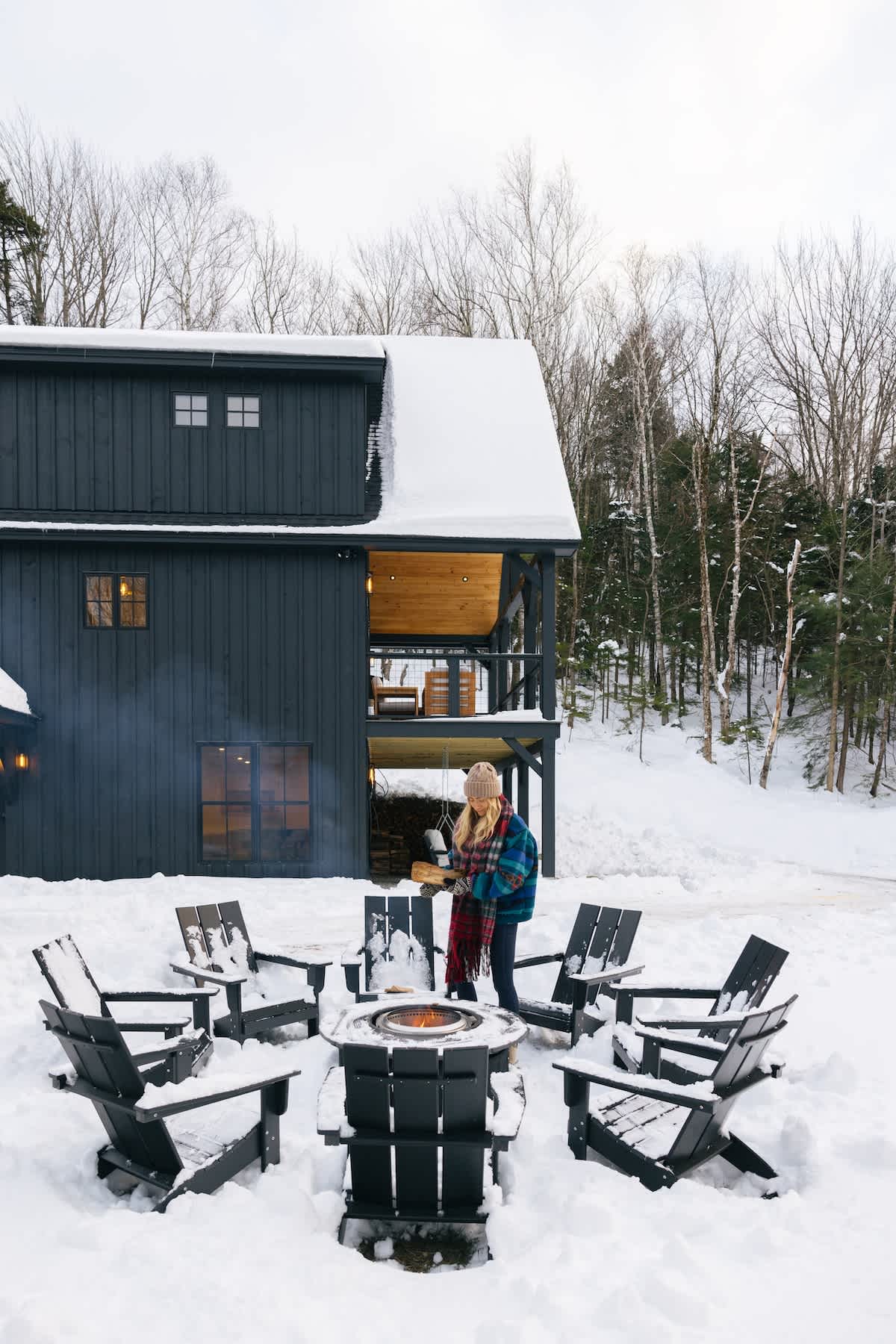 Daytime view of an outdoor fire pit area surrounded by dark Adirondack chairs, all covered in snow. A person in a plaid jacket tends to the fire, with the dark modern house and bare winter trees in the background under a cloudy sky.
