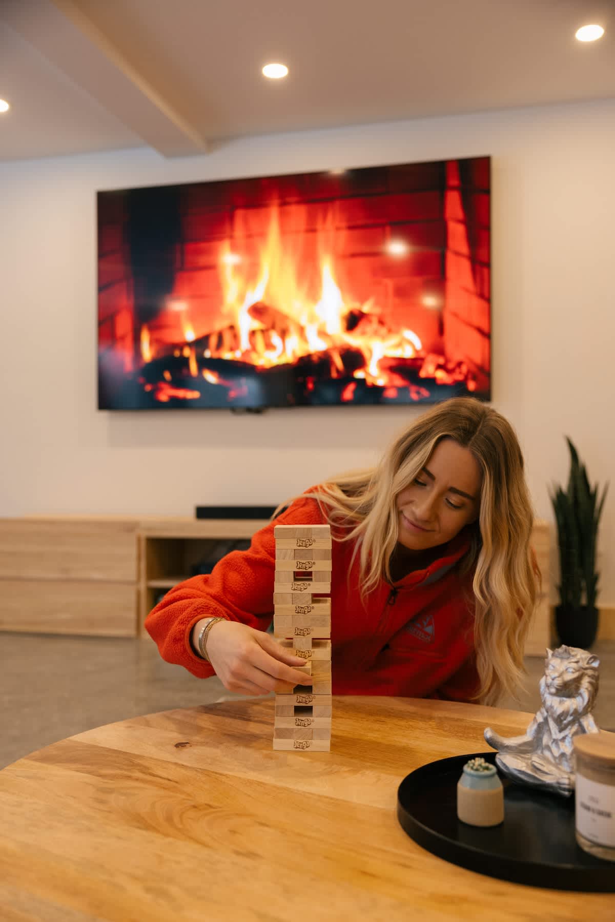 Bright recreation space with a blonde woman playing Jenga at a round wooden table, set against a large wall-mounted TV displaying a cozy fireplace scene. A low wooden console and a plant are visible in the background.
