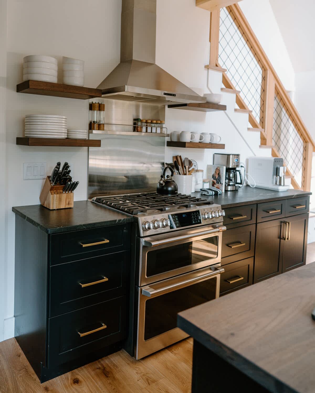 Modern kitchen featuring a stainless steel gas range with a double oven and matching hood, black countertops, and dark cabinetry with gold hardware. Open wooden shelves display white dishes and mugs, with a staircase visible in the background.