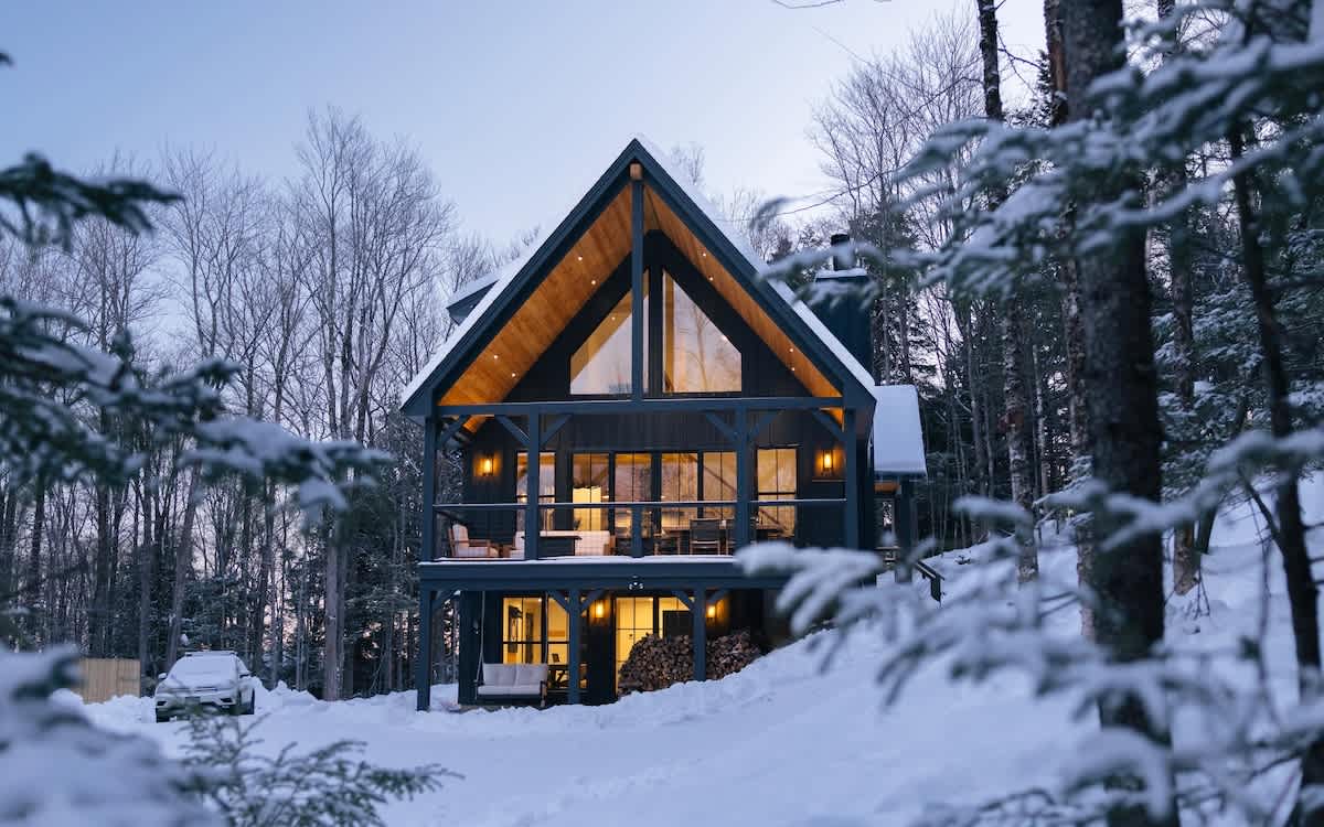 A striking A-frame cabin with dark siding and a prominent glass facade stands illuminated in a snow-covered forest at dusk. The multi-level structure features a balcony and warm interior lighting, creating a cozy winter retreat.
