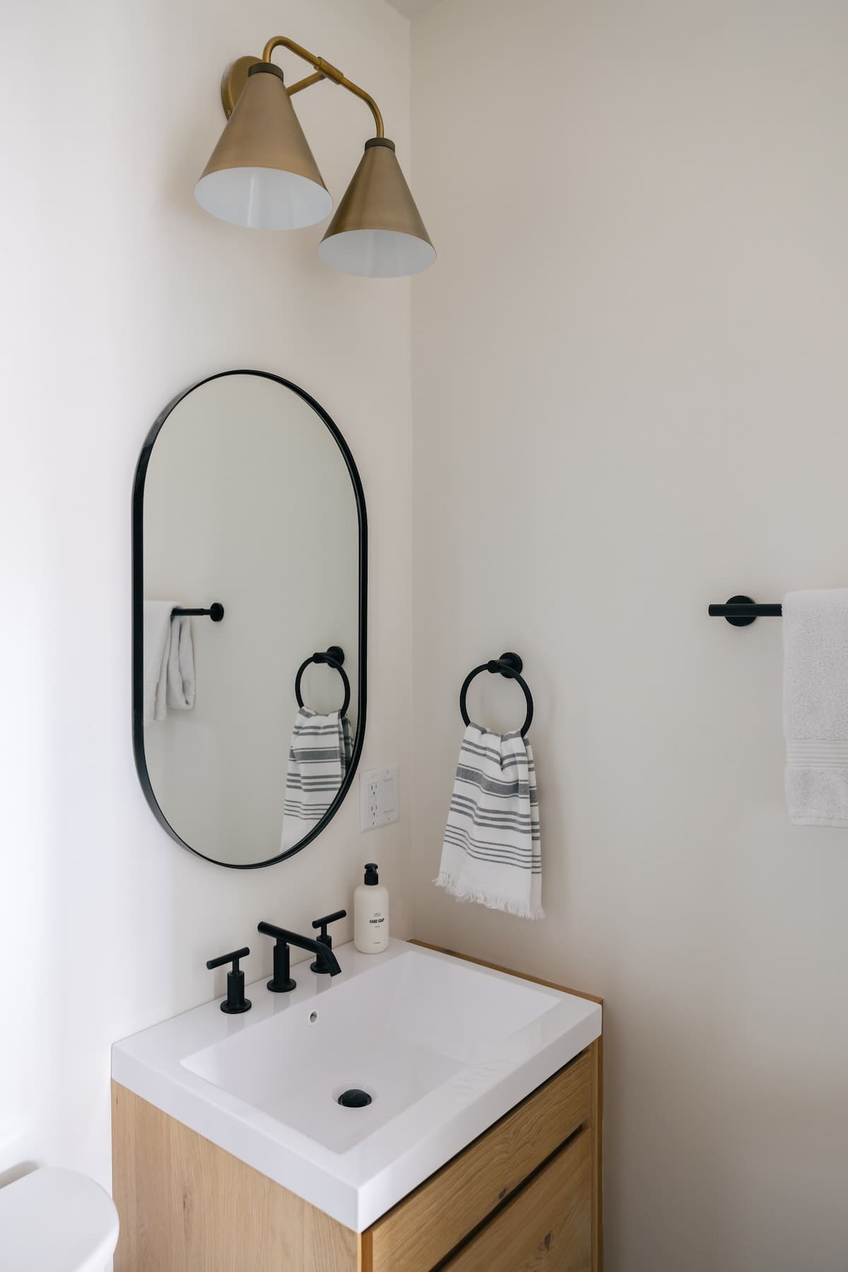 Modern bathroom with a white vanity, a black oval mirror, and black fixtures. A brass-colored double sconce light fixture is mounted above the mirror, and white towels hang on black towel rings.