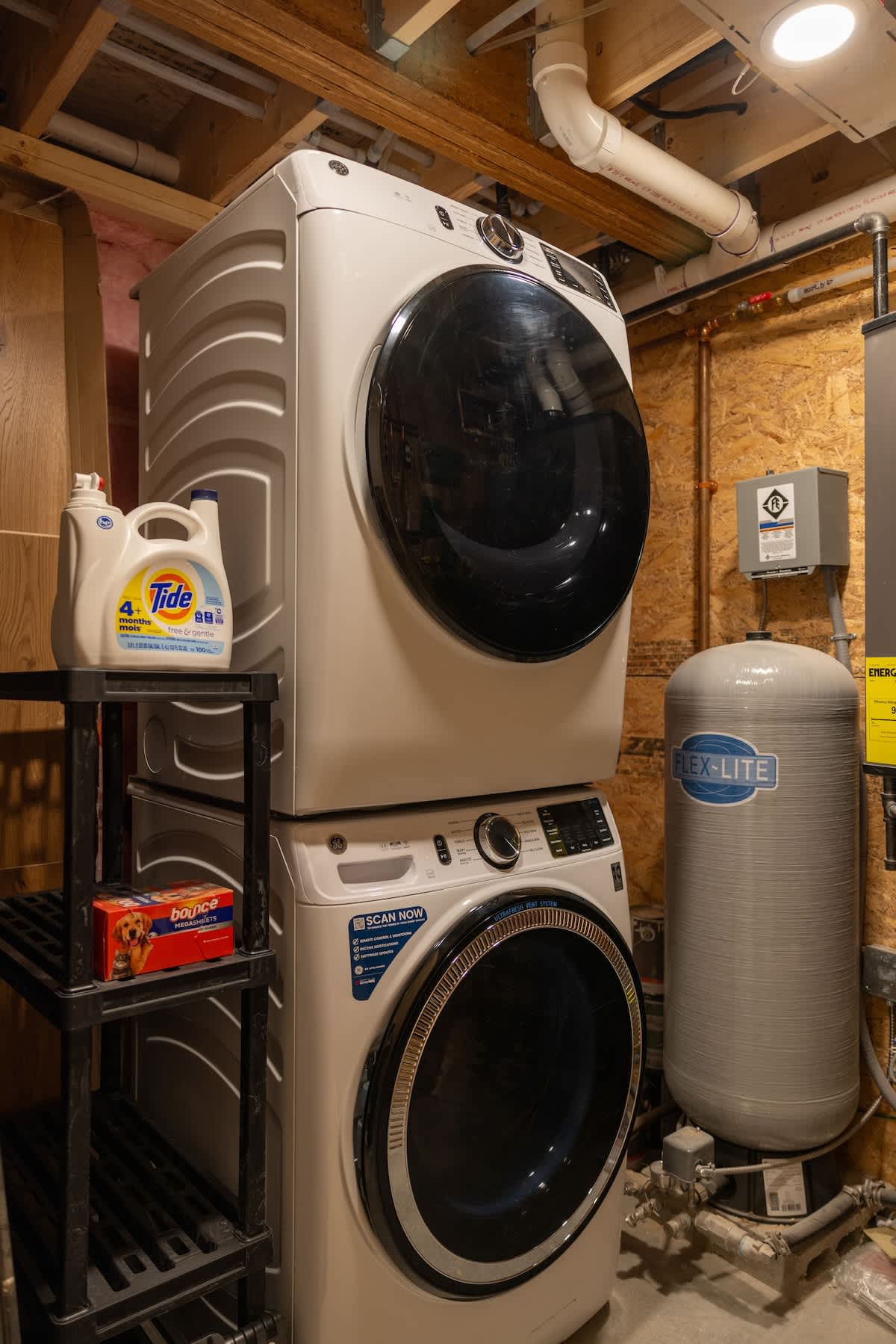 Functional laundry area featuring a stacked white washer and dryer unit, with laundry detergent and dryer sheets on a black shelving unit beside it. Exposed pipes and wooden walls indicate a utility space.