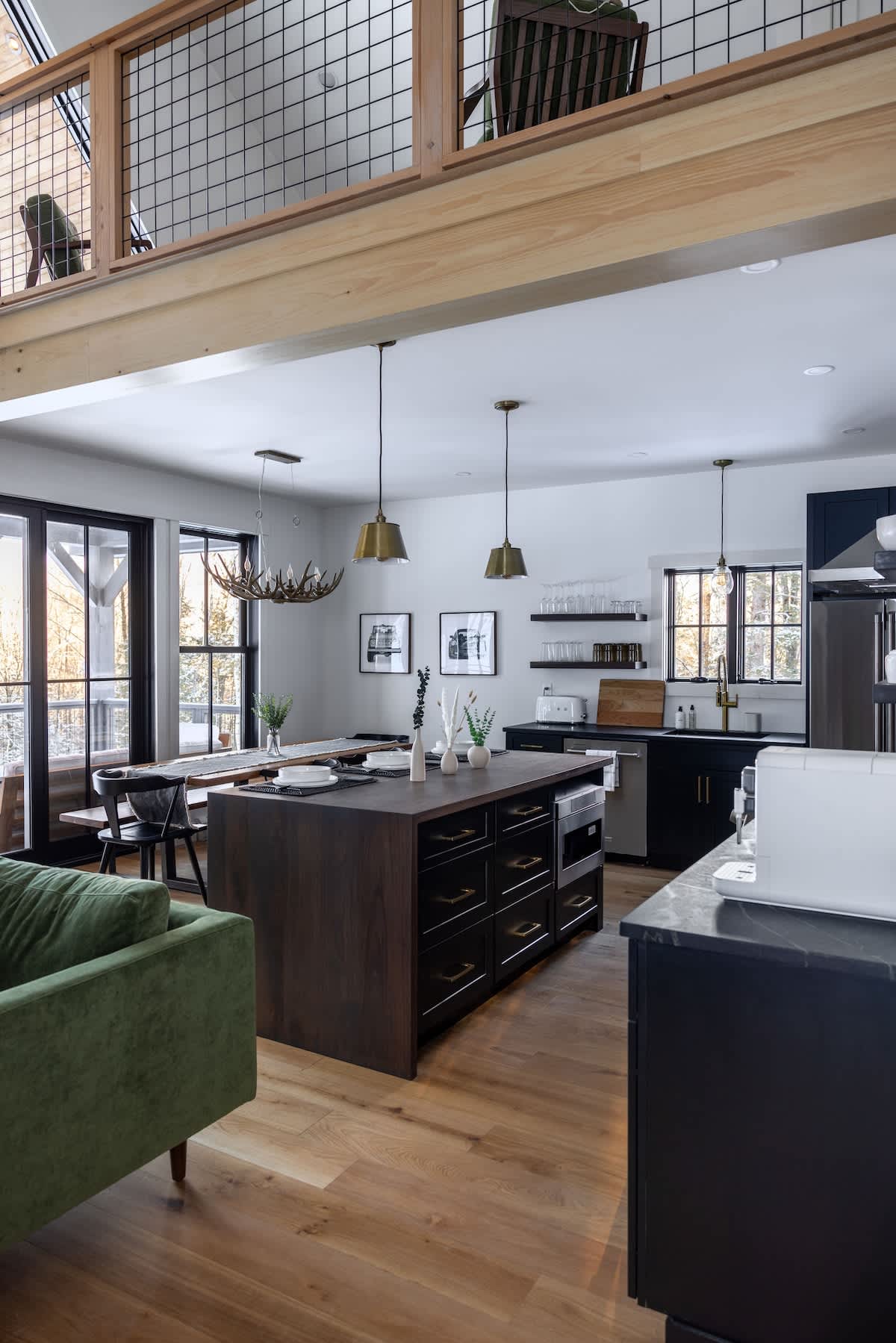 Modern open-concept kitchen and dining area featuring a dark wood island with an integrated microwave, brass pendant lights, and light hardwood floors. A long dining table with a unique antler chandelier sits next to large windows, while a green sofa is visible in the foreground.