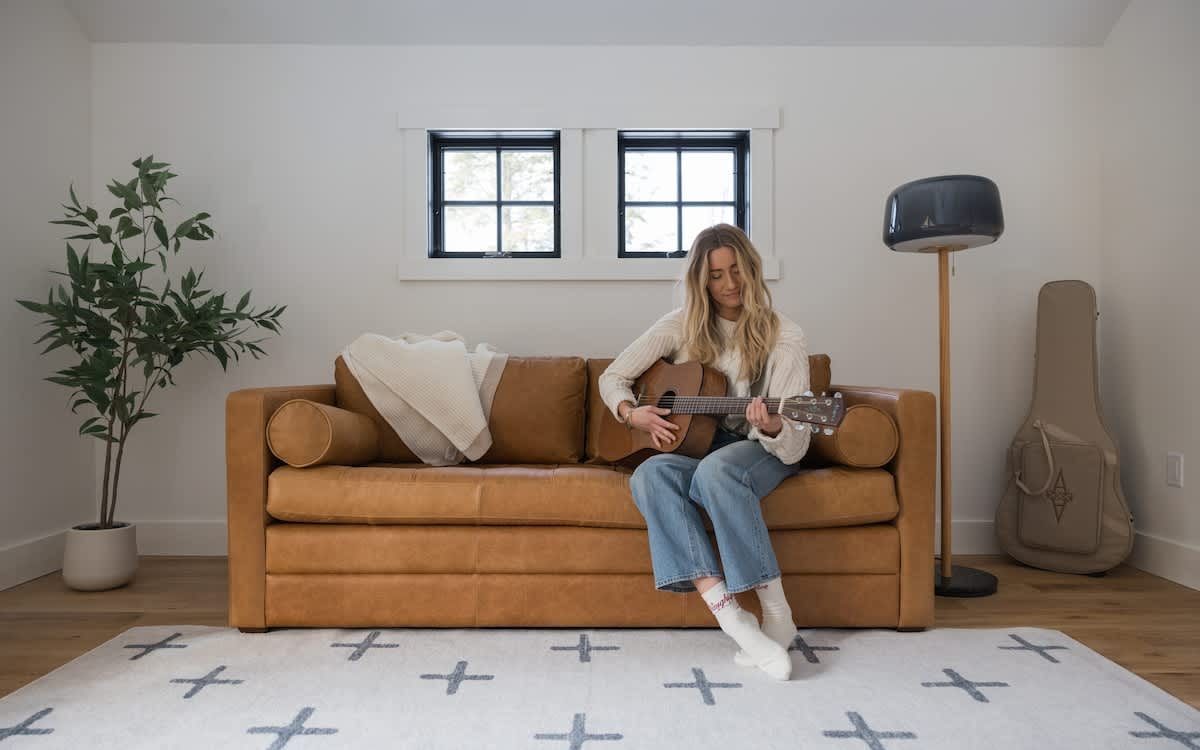 A woman plays an acoustic guitar while sitting on a stylish brown leather sofa in a bright living area. The room features light hardwood floors, a patterned rug, and a large potted plant, with two small windows above the sofa.