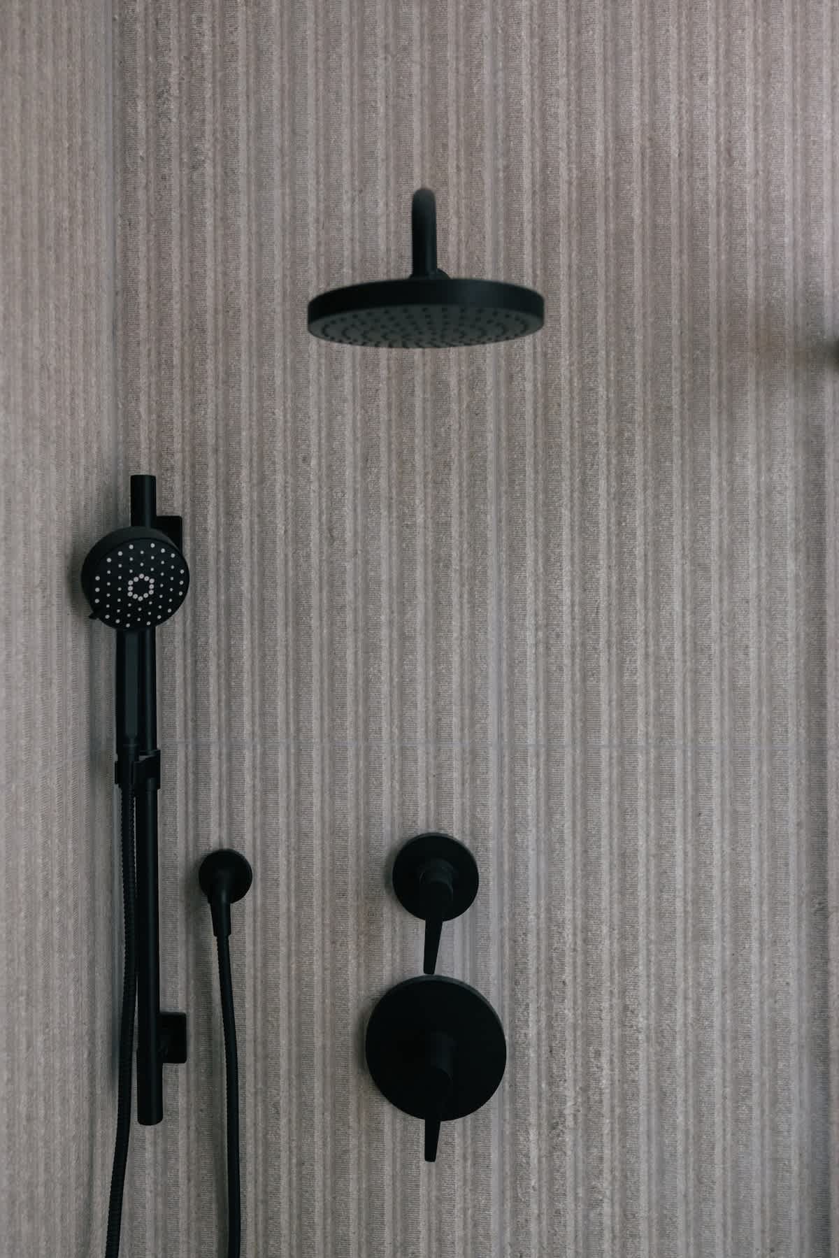 Close-up view of a modern shower with textured gray vertical stripe tiles, featuring a matte black rain shower head, a handheld shower, and minimalist black control knobs.