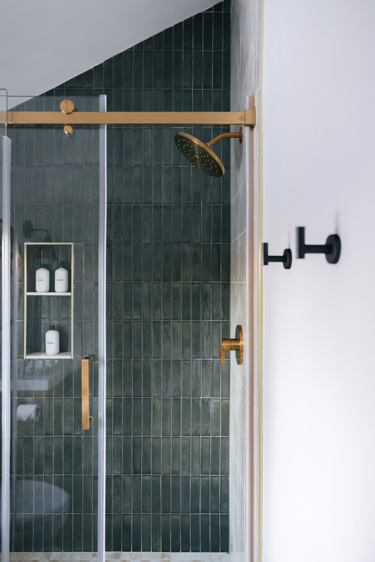 Modern shower stall with dark green subway tiles, a gold-framed glass door, and matching gold shower fixtures including a rain shower head. A built-in niche holds white bottles, and black wall hooks are visible outside the shower.