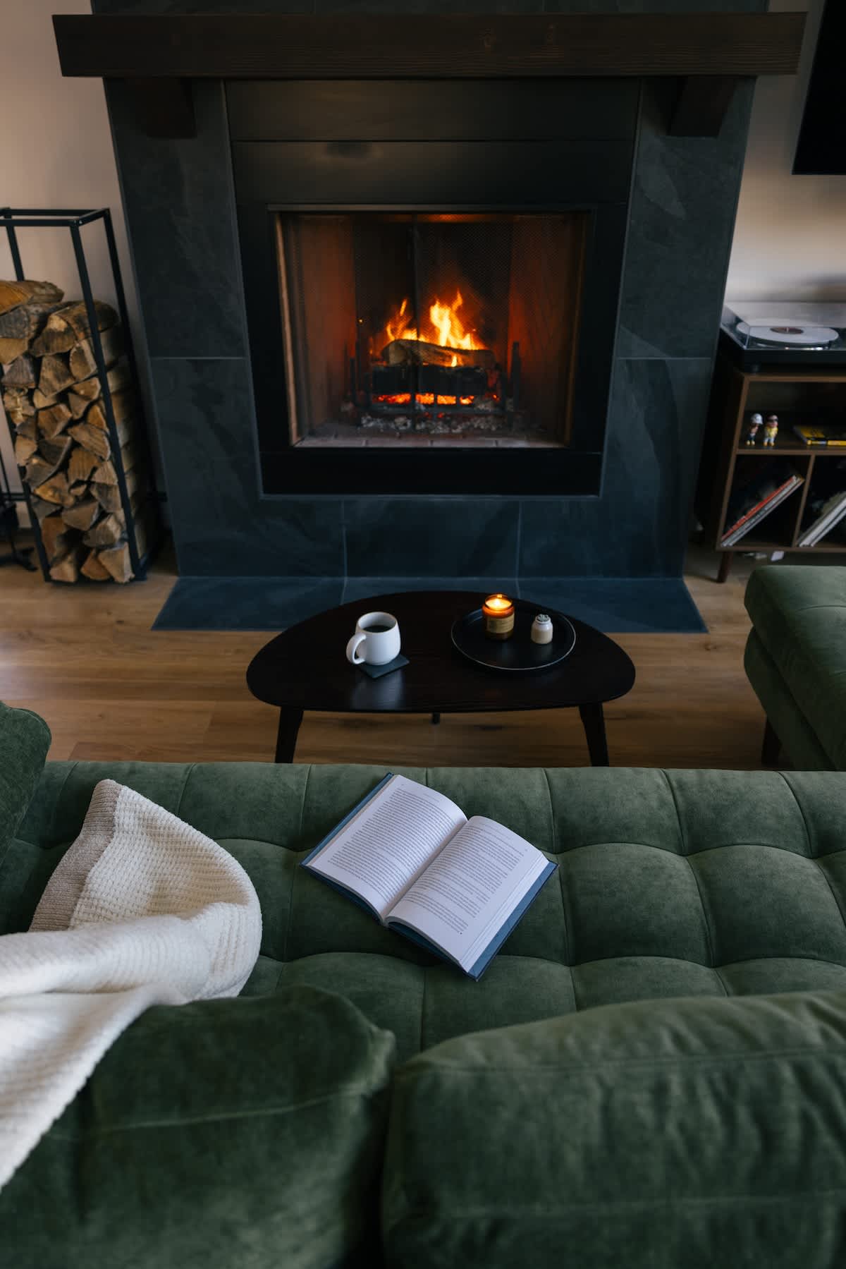 Cozy living room featuring a dark green tufted sofa with a white throw, a black coffee table with a mug and candle, and a modern fireplace with a dark stone surround and a roaring fire. Wood floors and a stacked woodpile are visible in the background.