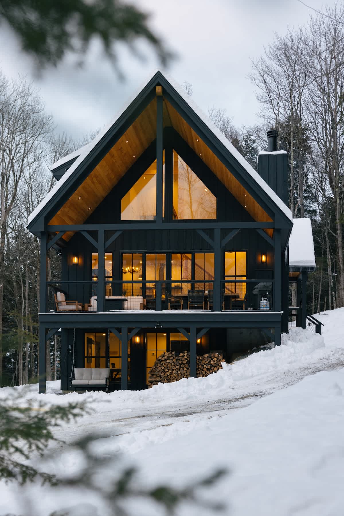 Grand A-frame modern cabin with a dark exterior and a prominent gabled roof, nestled among snow-covered trees. Large windows illuminate the interior, and a spacious deck with outdoor seating extends from the main level, with firewood stacked below.