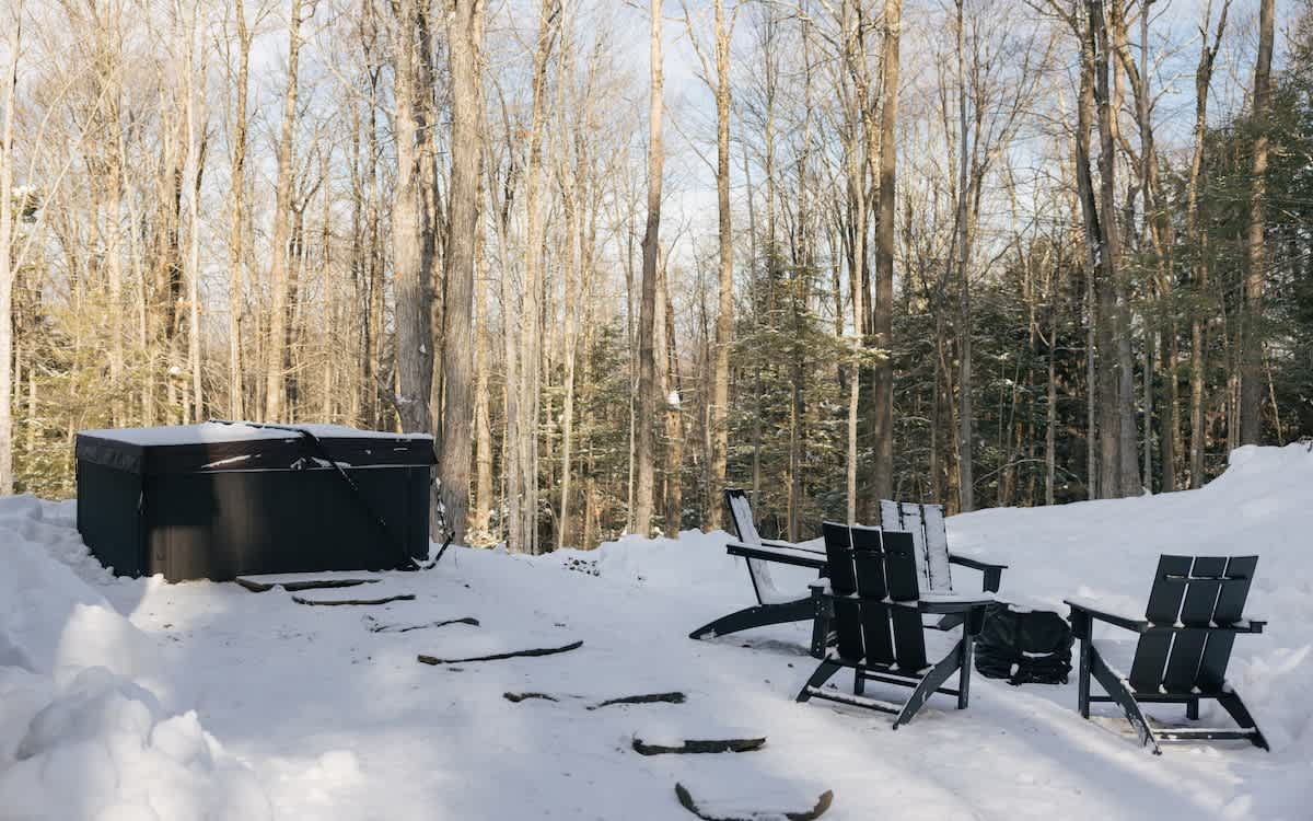A hot tub is situated in a snowy backyard surrounded by tall, bare trees, with several black Adirondack chairs and a fire pit nearby. Stone steps lead up to the hot tub area, creating a serene winter outdoor space.