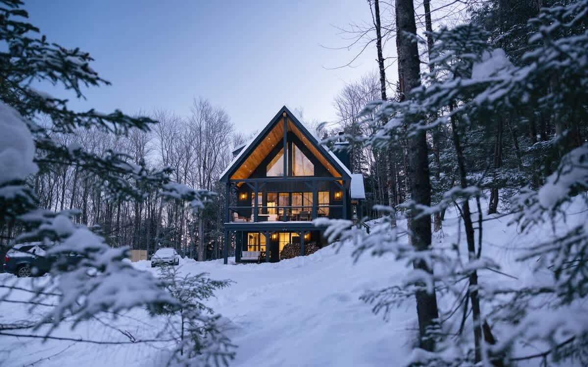 A modern A-frame house with large glass windows and warm interior lighting stands majestically in a snow-covered forest at dusk. Snow-laden evergreen branches frame the view of the inviting cabin.