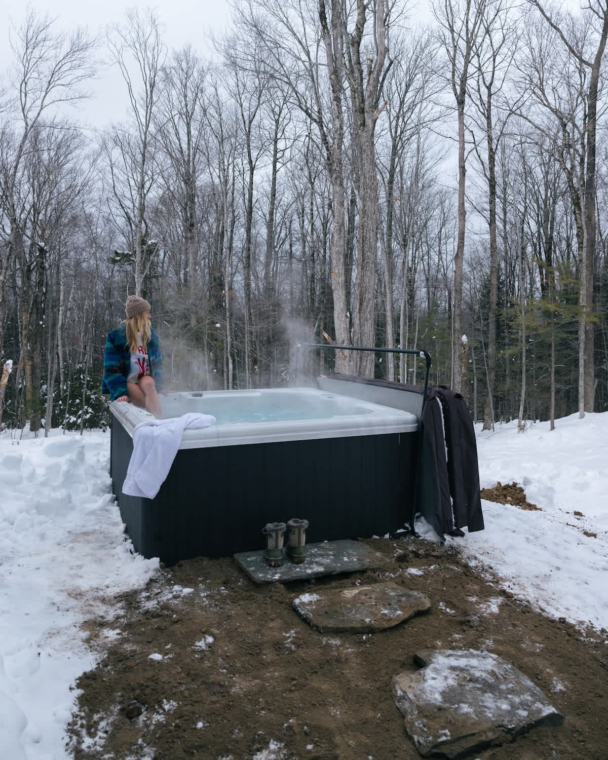A person relaxes in a steaming outdoor hot tub, surrounded by a snow-covered forest with bare trees under a cloudy sky. A white towel hangs over the tub's edge, and stone steps lead up to it.