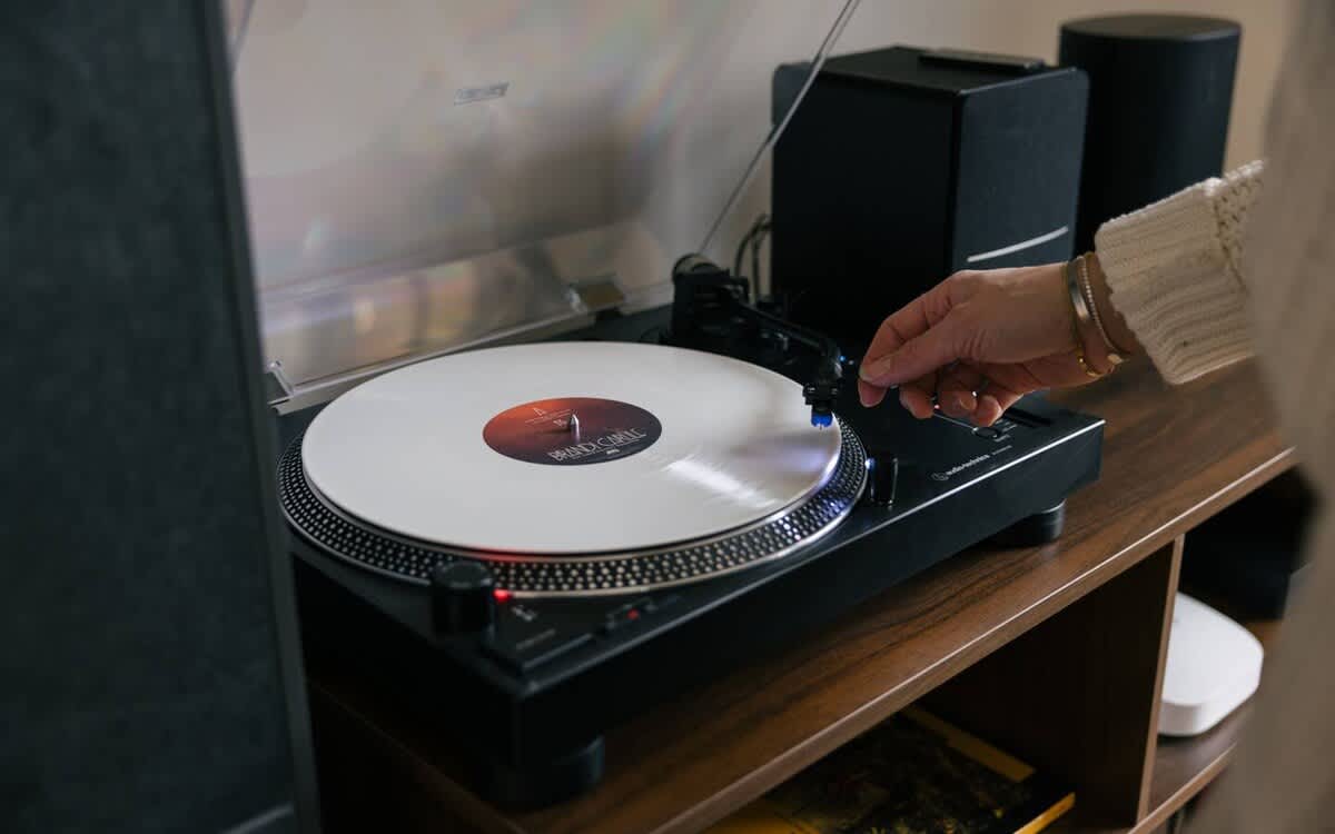 A close-up shot of a hand placing a needle on a white vinyl record playing on a modern turntable. The turntable is set on a wooden media console, flanked by black speakers.