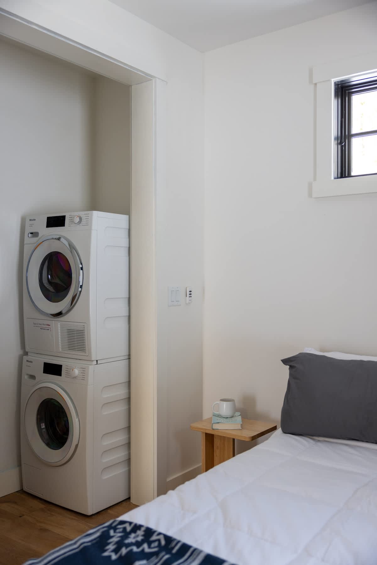 A stacked white washer and dryer unit is neatly integrated into a closet alcove within a bedroom, adjacent to a bed with white bedding and a gray pillow. A small wooden nightstand sits beside the bed.