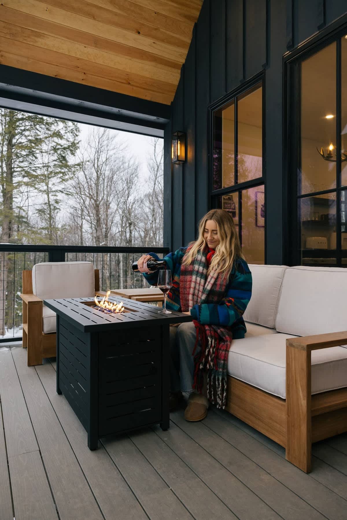 A woman in a plaid blanket scarf pours wine on a covered outdoor deck with a modern black fire pit table. The deck features comfortable outdoor seating and overlooks a snowy, wooded landscape.