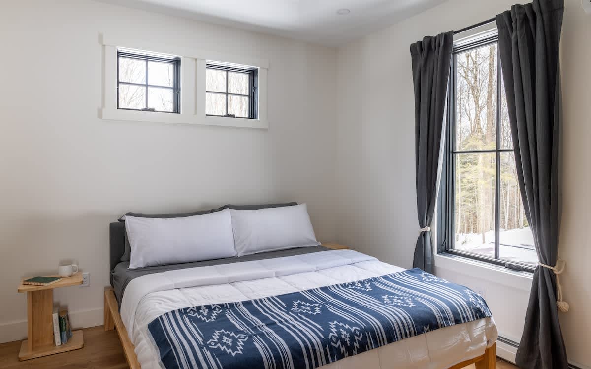 Cozy bedroom with a queen-sized bed featuring white and blue bedding, a light wood nightstand, and two large windows with dark gray curtains overlooking a snowy outdoor scene. Two smaller transom windows are positioned above the bed.