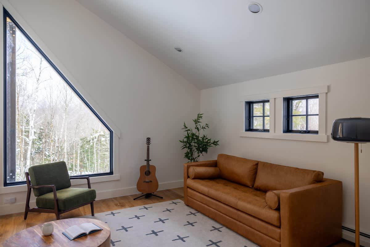 Bright living area with a tan leather sofa, a green armchair, and a wooden coffee table on a patterned rug. Large, angular windows offer views of a snowy forest, and a guitar stands in the corner.