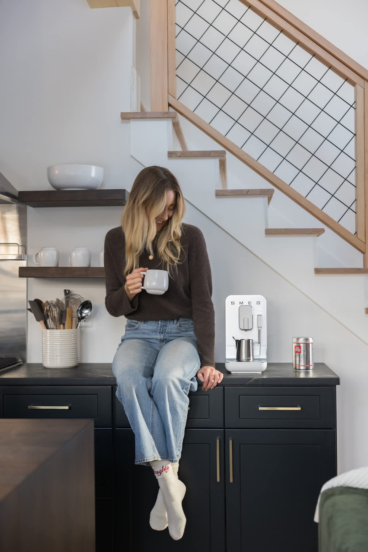Modern kitchen featuring dark cabinetry with gold hardware and a white countertop, where a woman sits holding a coffee mug. A white espresso machine and a staircase with a wire mesh railing are visible in the background.