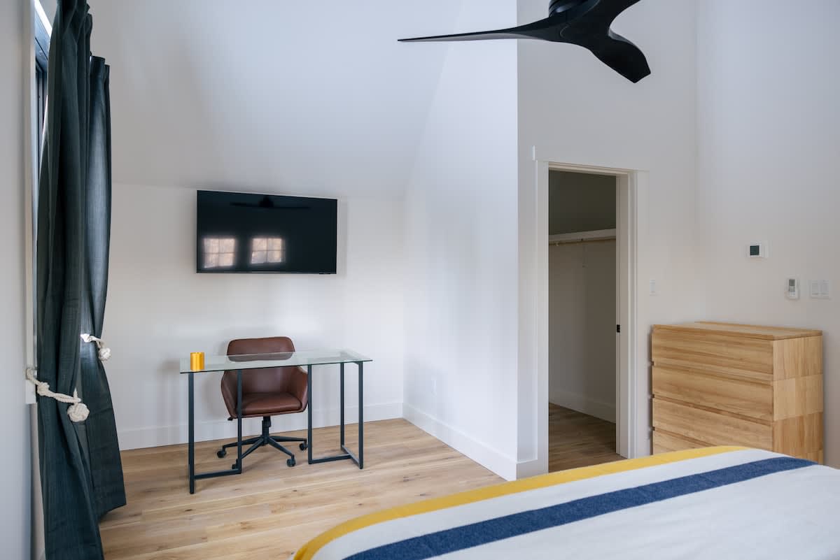Bright bedroom with light hardwood floors and white walls, featuring a wall-mounted TV above a glass-top desk with a brown office chair. A modern black ceiling fan hangs overhead, and a wooden dresser is visible next to an open closet door.