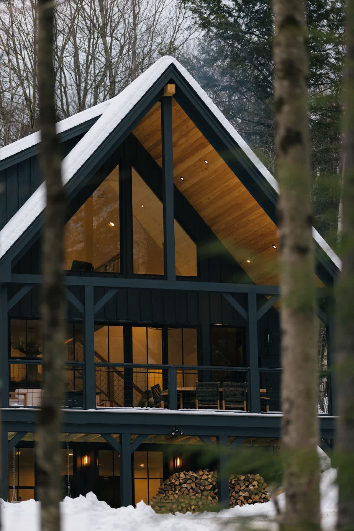 Close-up view of the modern A-frame cabin's front facade, showcasing large triangular windows and a covered deck with outdoor seating. The dark exterior contrasts with the snow-covered ground and surrounding bare trees.