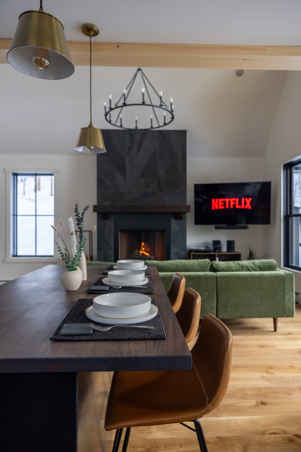 Open-concept living and dining area featuring a long dark wood dining table set with white dishes and brown leather bar stools, with the living room and fireplace visible in the background. Modern light fixtures hang above the table.