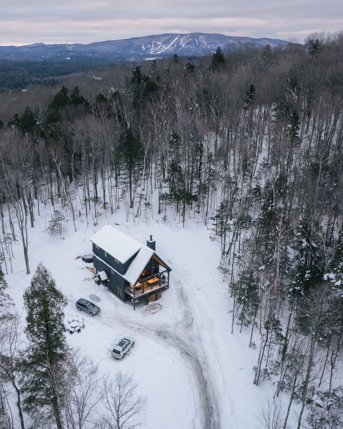 An aerial shot captures the dark-sided cabin surrounded by a snow-covered forest and a winding driveway, with two cars parked nearby. Distant mountains are visible under a cloudy sky, highlighting the secluded winter setting.