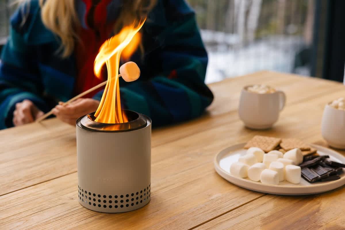 A person roasts a marshmallow over a small, modern tabletop fire pit on a light wooden table. S'mores ingredients (marshmallows, chocolate, graham crackers) are neatly arranged on a white plate beside it, with mugs in the background.