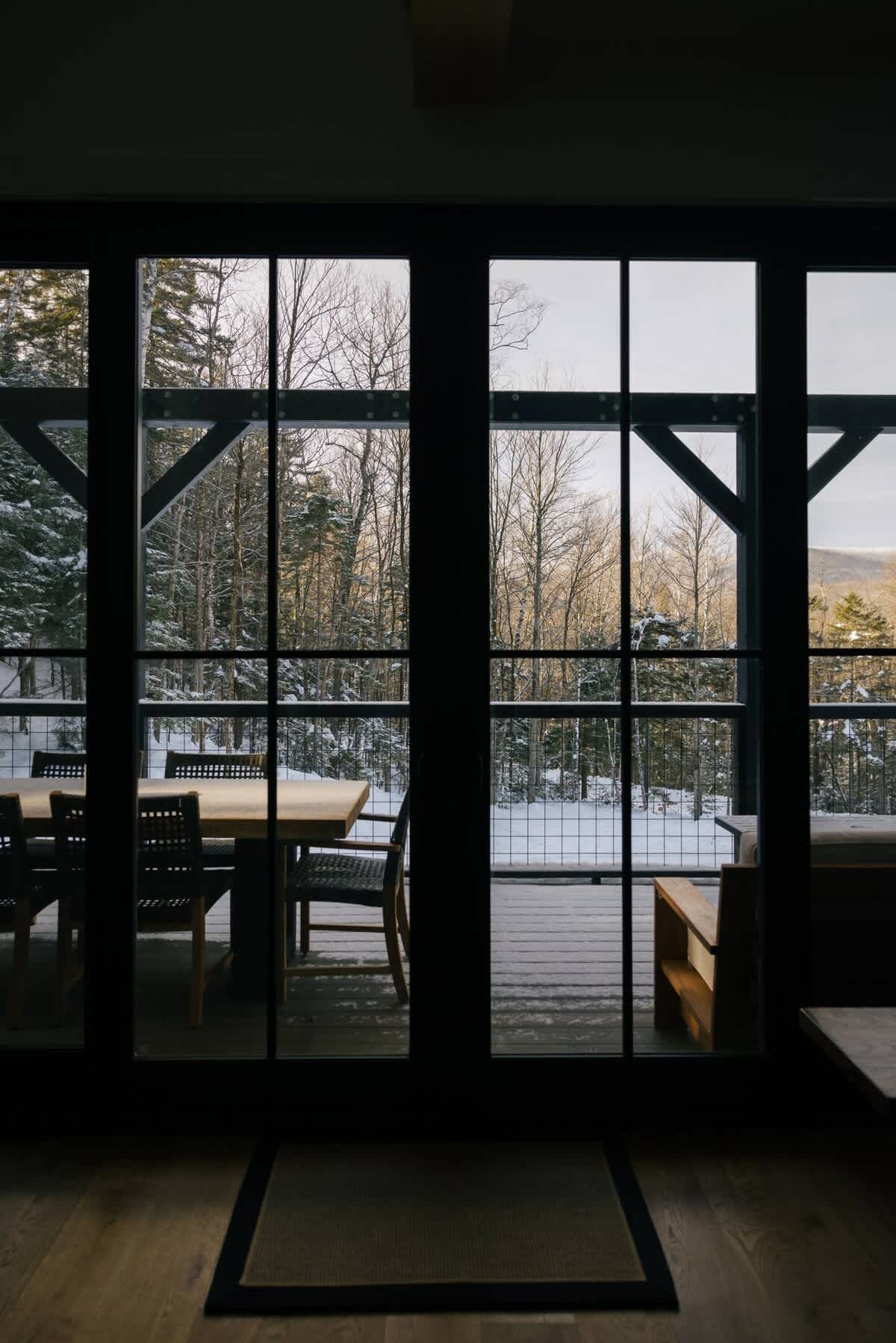 A view from inside through large black-framed glass doors reveals an outdoor deck furnished with a dining table and chairs, overlooking a serene snow-covered forest. The interior floor is light wood.