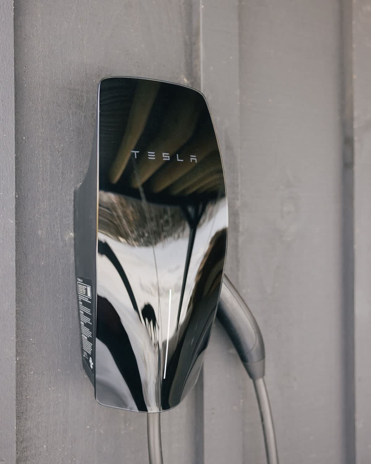 A sleek black Tesla wall connector is mounted on a dark gray wall, reflecting the surrounding environment. The charging cable is neatly coiled and hangs below the unit.