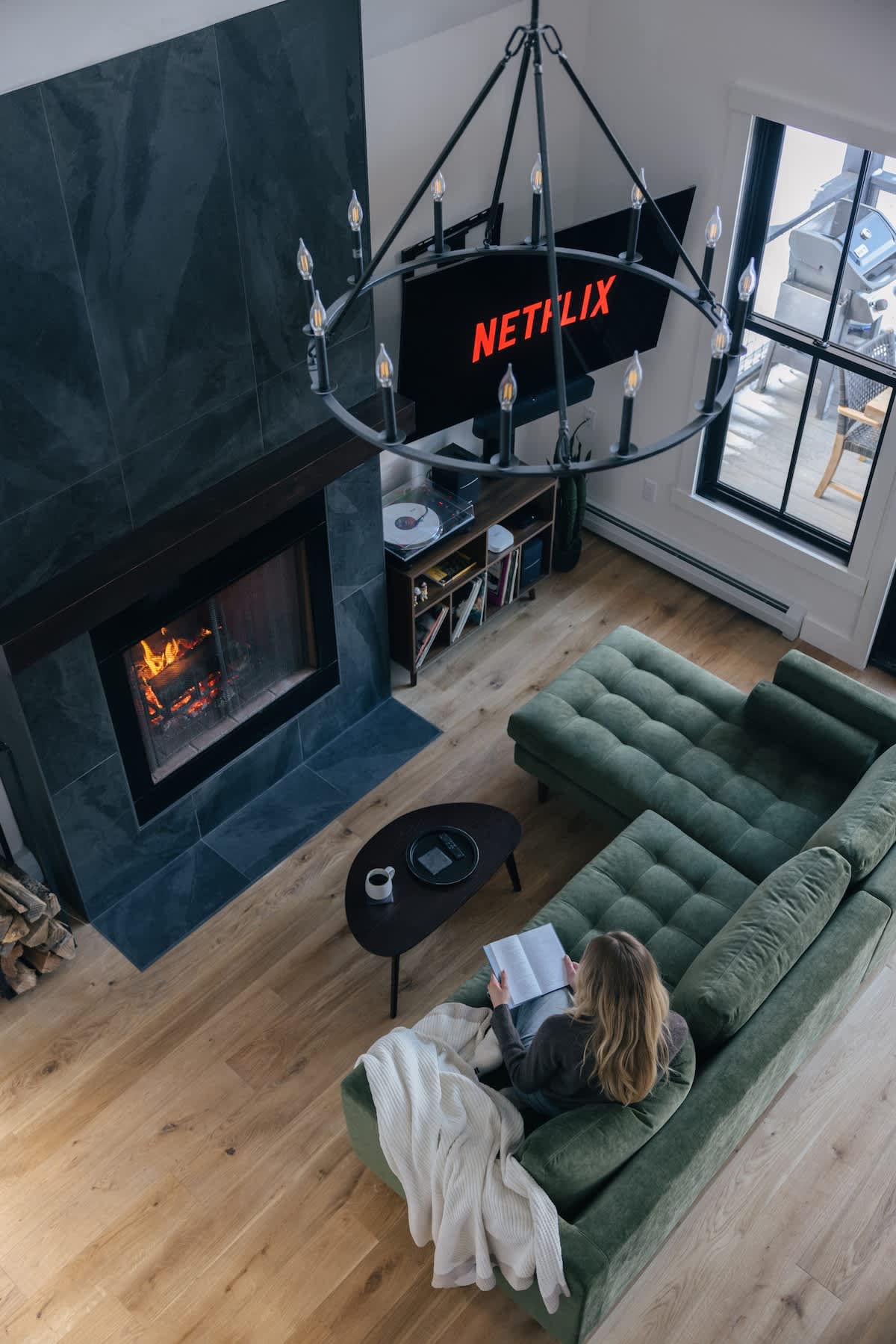 An aerial view of the living room shows a person reading on a green velvet sectional sofa facing a large TV mounted above a dark stone fireplace with a roaring fire. A large black chandelier hangs overhead, and hardwood floors extend throughout the space.
