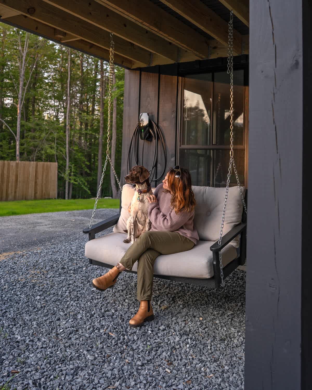 Inviting outdoor porch swing with a woman and a dog relaxing on it, set against a backdrop of a gravel driveway and a wooded area. The swing is under a covered porch with dark siding.