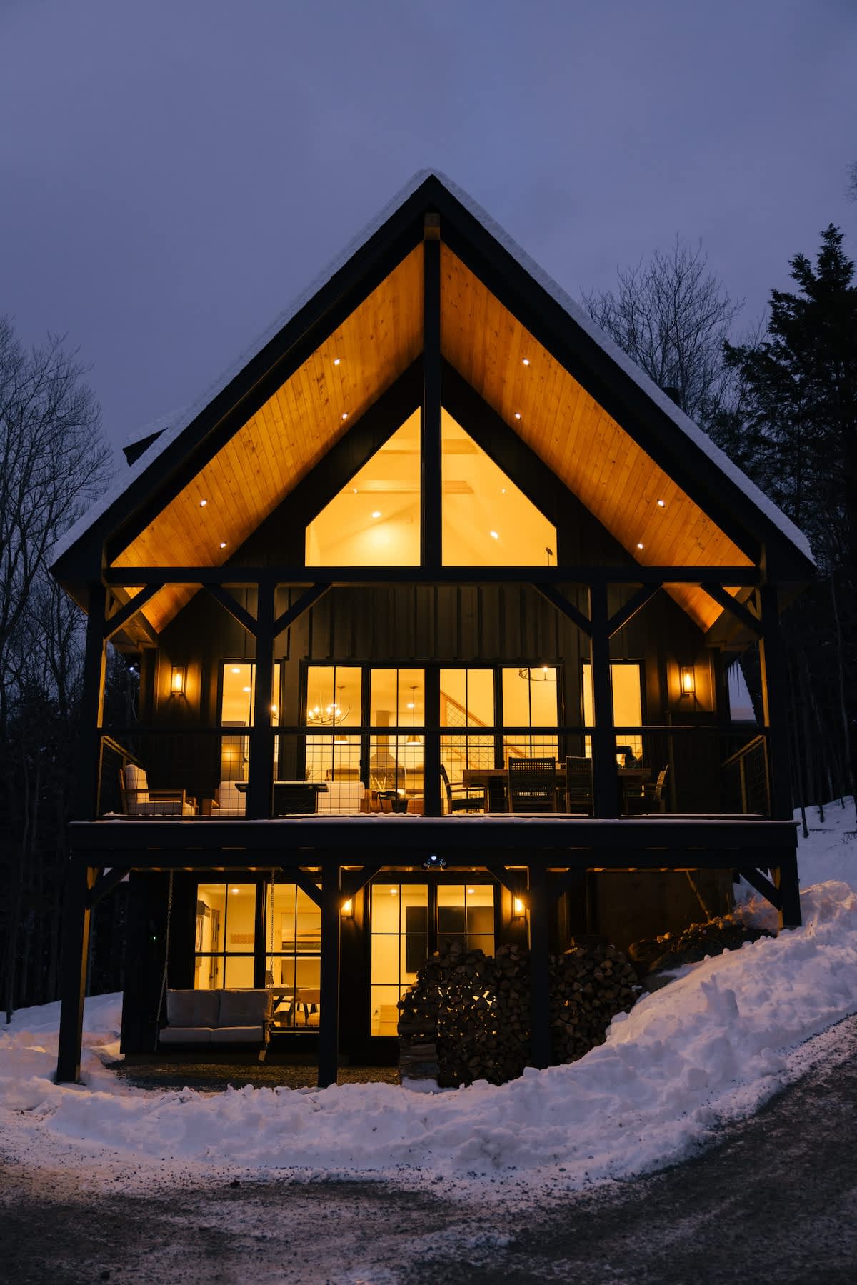 Striking A-frame modern cabin at dusk, featuring a dark exterior with warm lights glowing from large windows and a vaulted ceiling. A spacious deck extends across the front, overlooking a snow-covered landscape and bare winter trees.