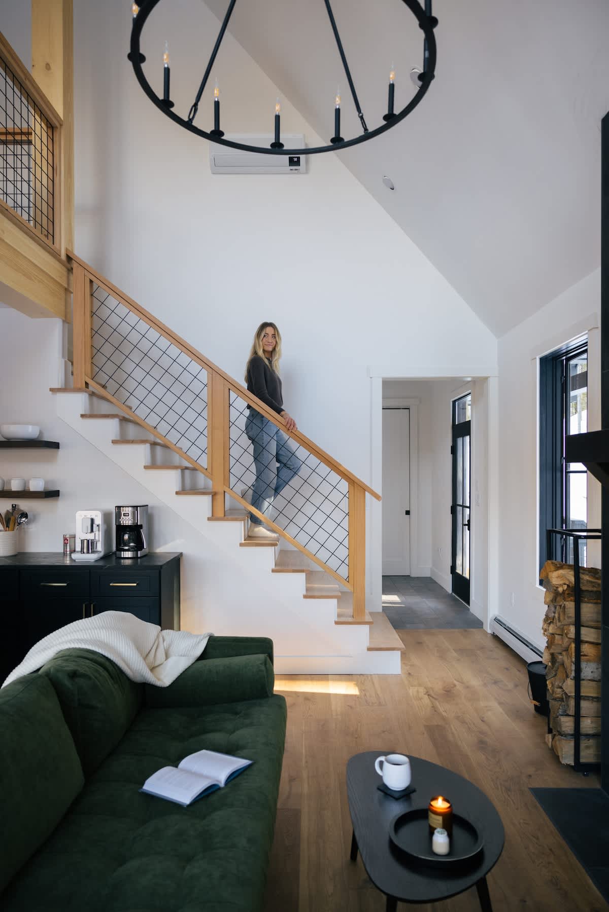 A woman descends a modern wooden staircase with a black metal mesh railing, leading into an open-concept living area with a green sofa and a fireplace. The space features high ceilings and hardwood floors.