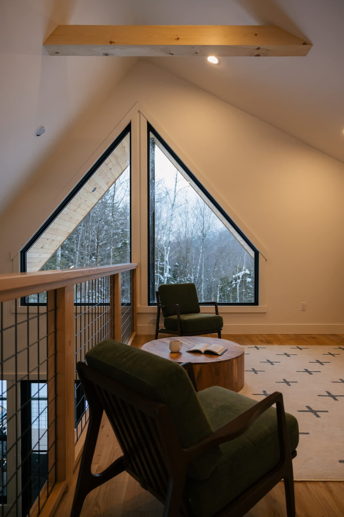 Elevated loft lounge area with green armchairs and a wooden coffee table, offering expansive views of a snowy forest through large triangular windows. The space features hardwood floors and a vaulted ceiling.
