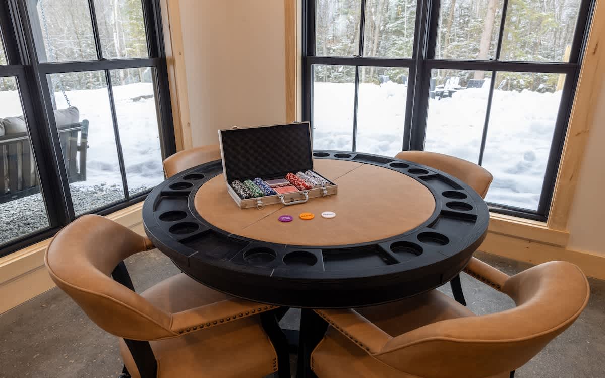 A round poker table with a tan leather surface and black chip holders is set with a metal case of poker chips and colorful playing chips. Four tan leather chairs surround the table, with large windows offering snowy forest views.