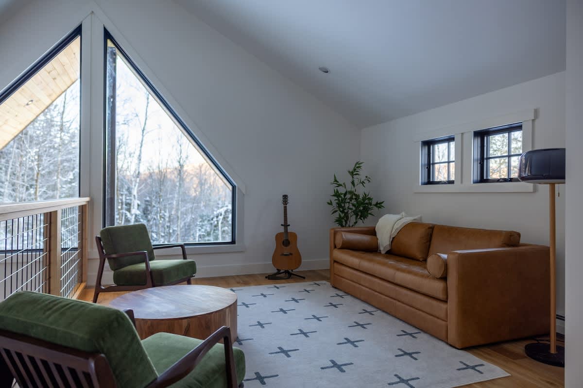 An upstairs loft living area features a brown leather sofa, two green armchairs, a round wooden coffee table, and a patterned rug. Large triangular windows offer panoramic views of a snowy, wooded landscape.