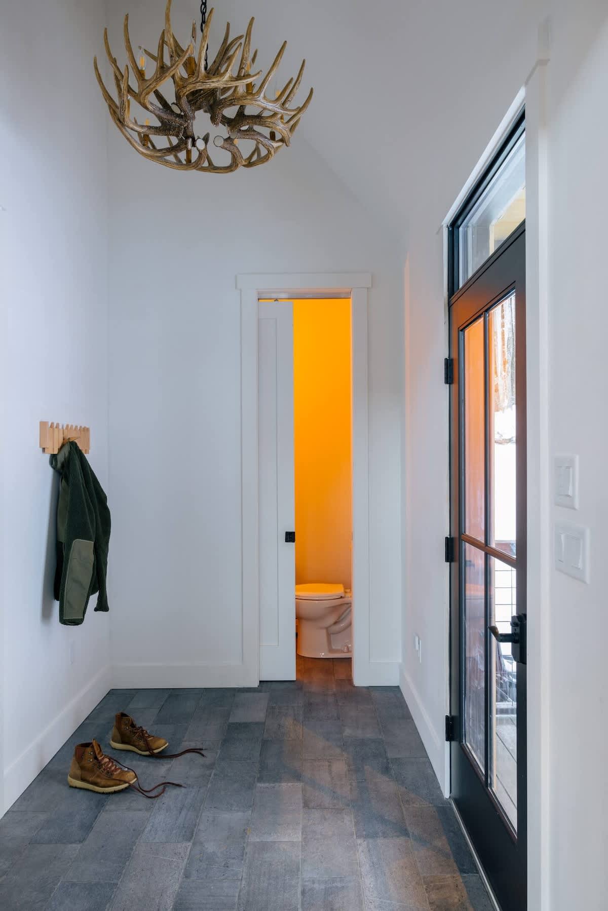 Bright entryway with dark gray herringbone tile flooring, a wooden coat rack with a jacket, and a pair of boots. A pocket door reveals a powder room with warm lighting, and a glass-paned exterior door is visible on the right.