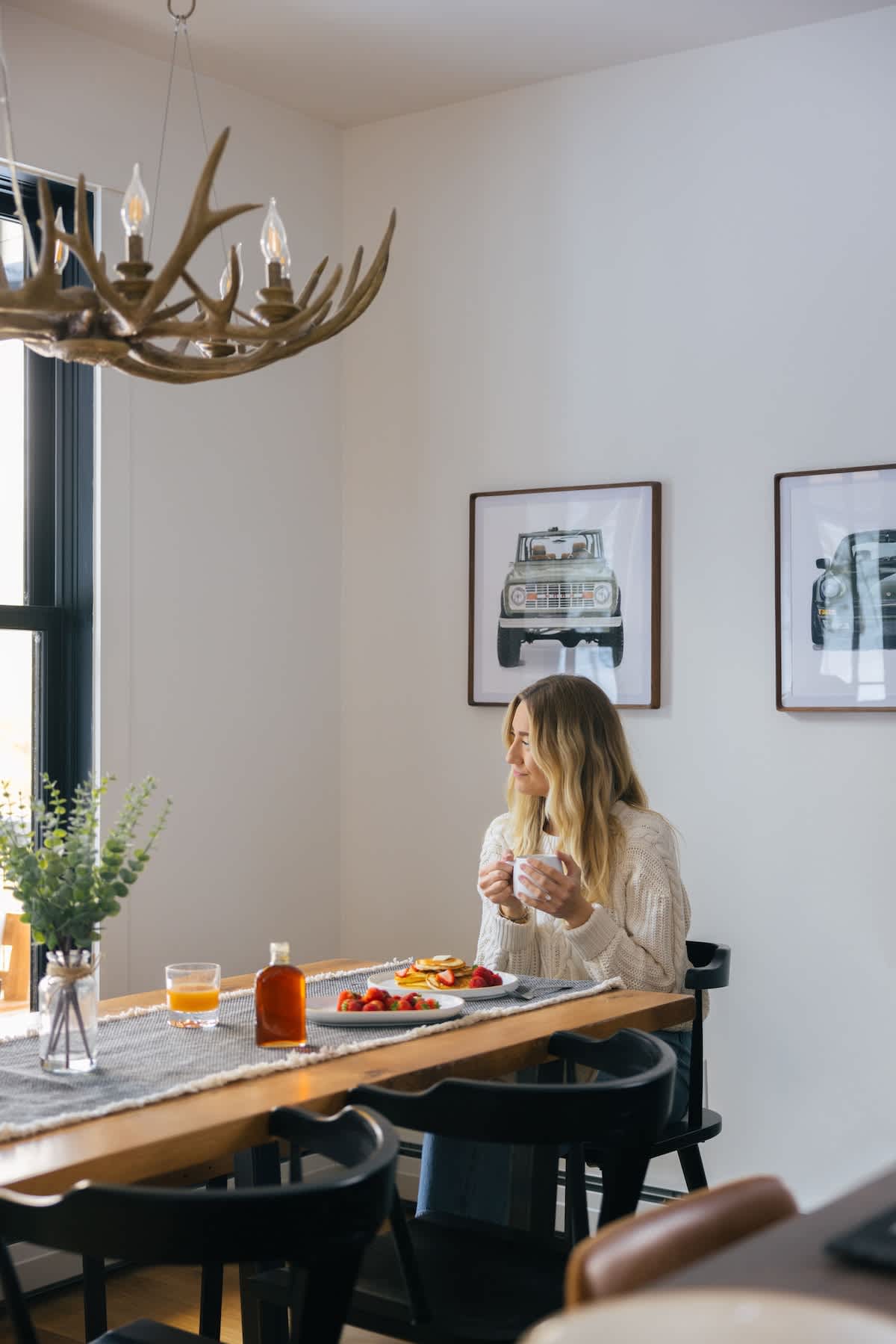A woman enjoys breakfast with pancakes and fruit at a long wooden dining table with black chairs, beneath an antler-style chandelier. The bright room has large windows and framed artwork on the wall.