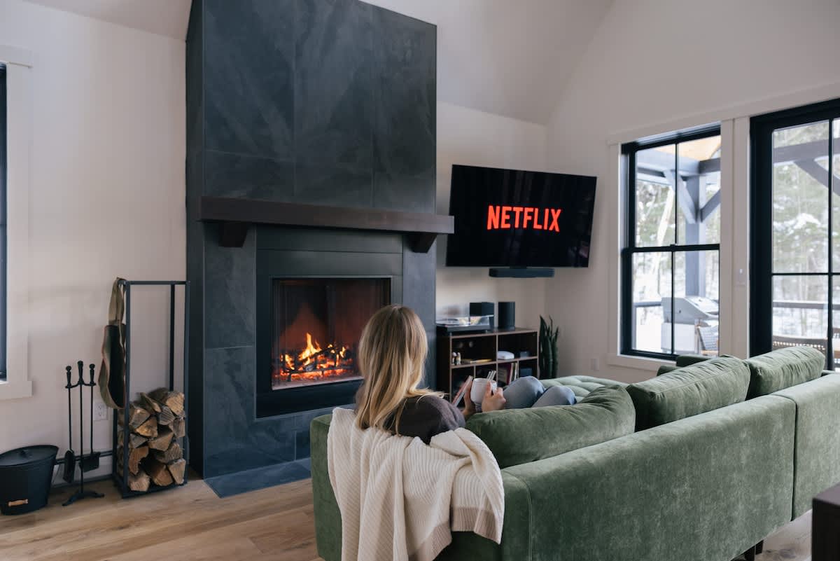 Spacious living room with a large green sectional sofa facing a modern black slate fireplace with a wooden mantel and a wall-mounted TV displaying Netflix. A person is relaxing on the sofa, enjoying the warmth of the fire.