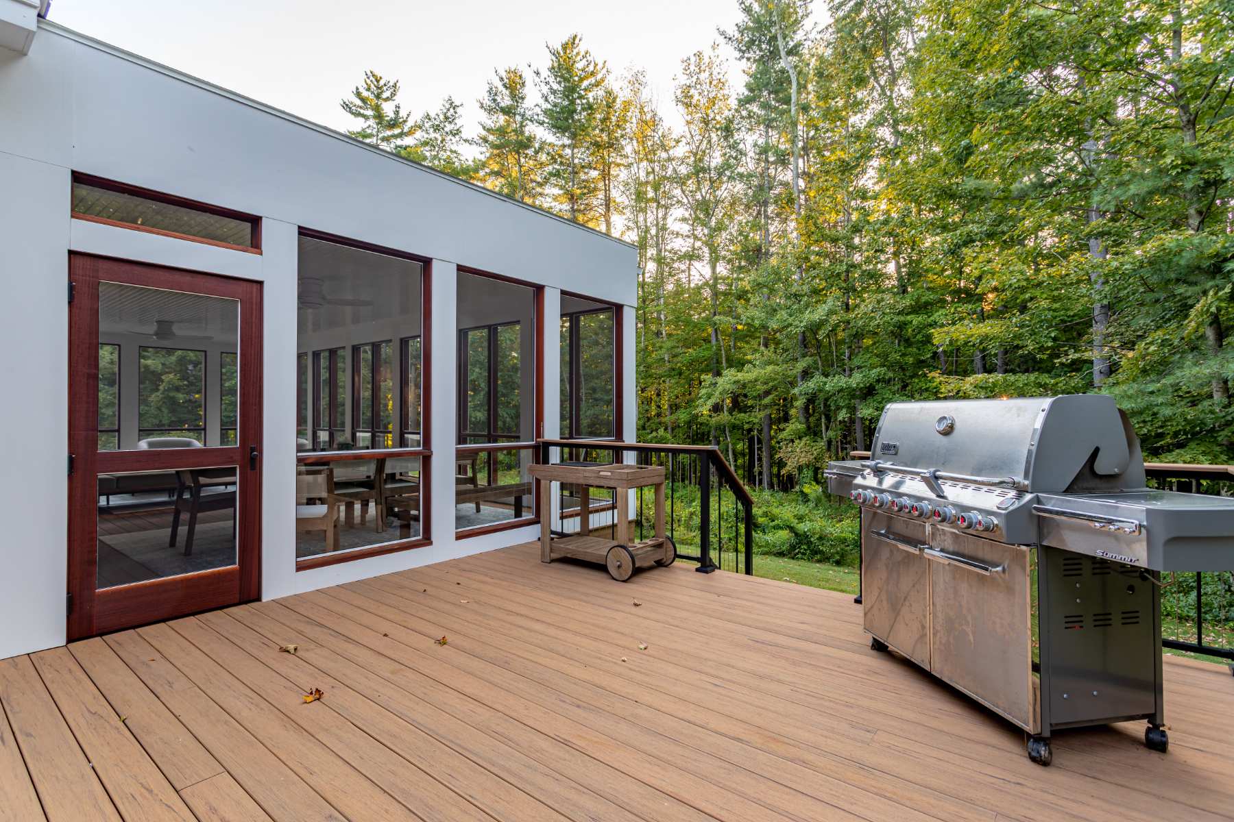 An expansive outdoor deck made of composite wood, featuring a large stainless steel BBQ grill and a screened-in porch area. The deck is surrounded by lush green trees, suggesting a private, natural setting.